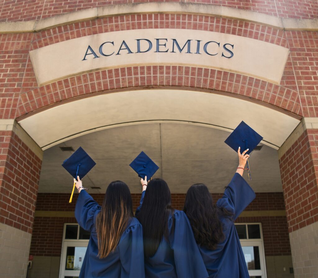 Three girls in blue graduation robes facing an Academics building holding up their caps.