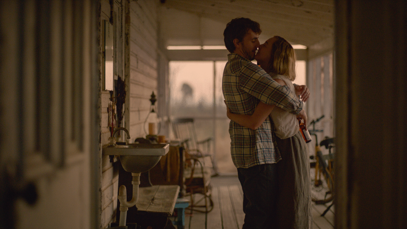 A scene of the movie FOE showing two people hugging and leaning for a kiss on the front porch of a house. 
